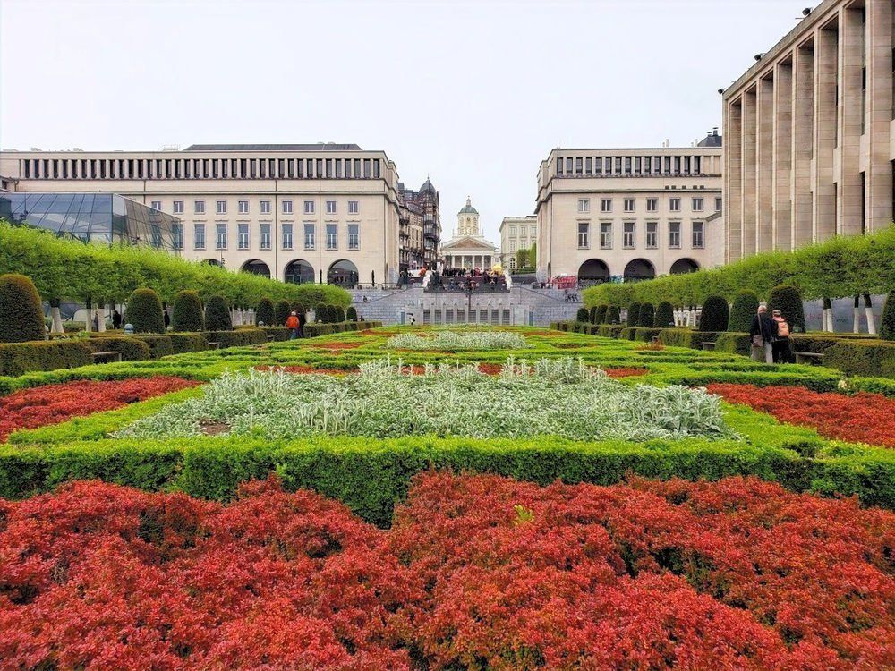 Formal garden with red, green, and white patterned hedges leading up to a building with columns, in Brussels.
