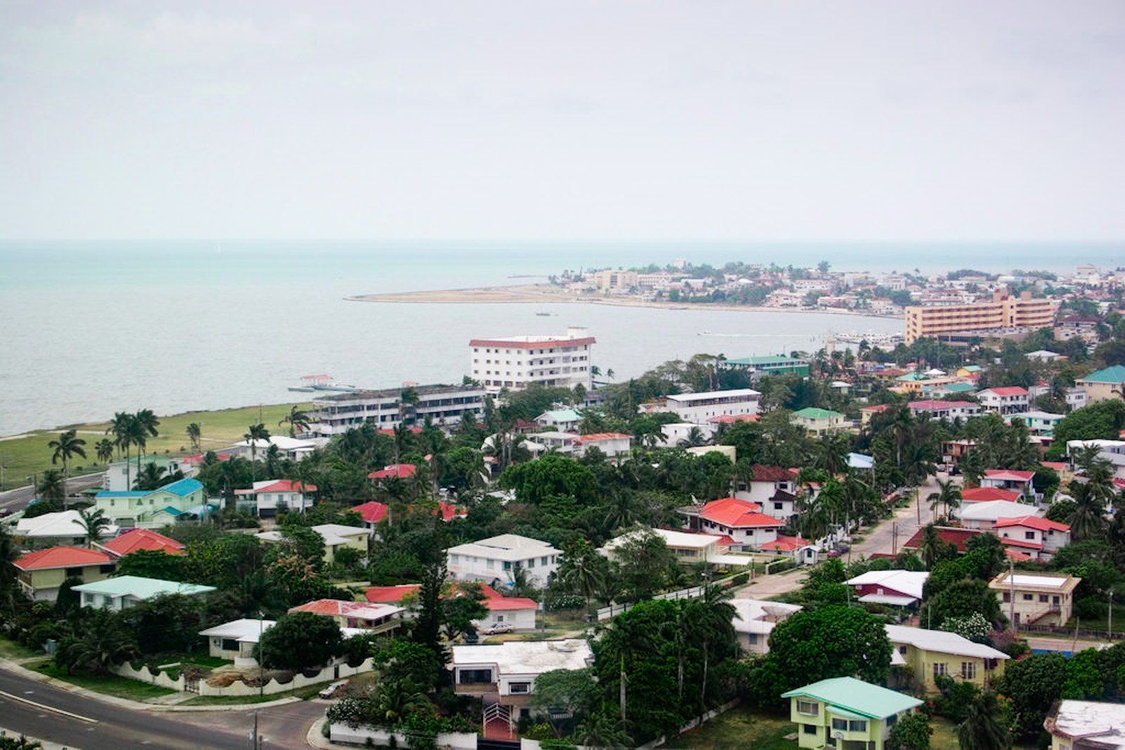 Coastal town view with buildings, lush trees, and the sea under a cloudy sky.