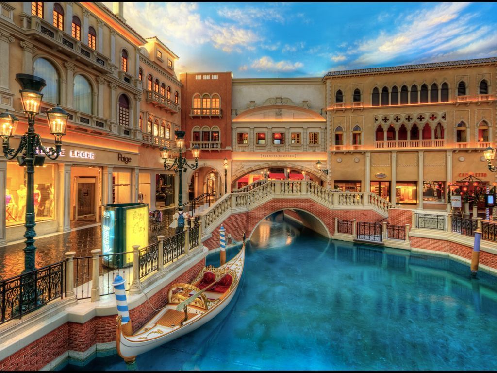 A gondola floats on blue water in a Venetian-themed setting with shops and bridges under a blue sky.