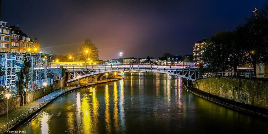 Nighttime view of a bridge over a river, with city lights reflecting on the water's surface and buildings on either side.