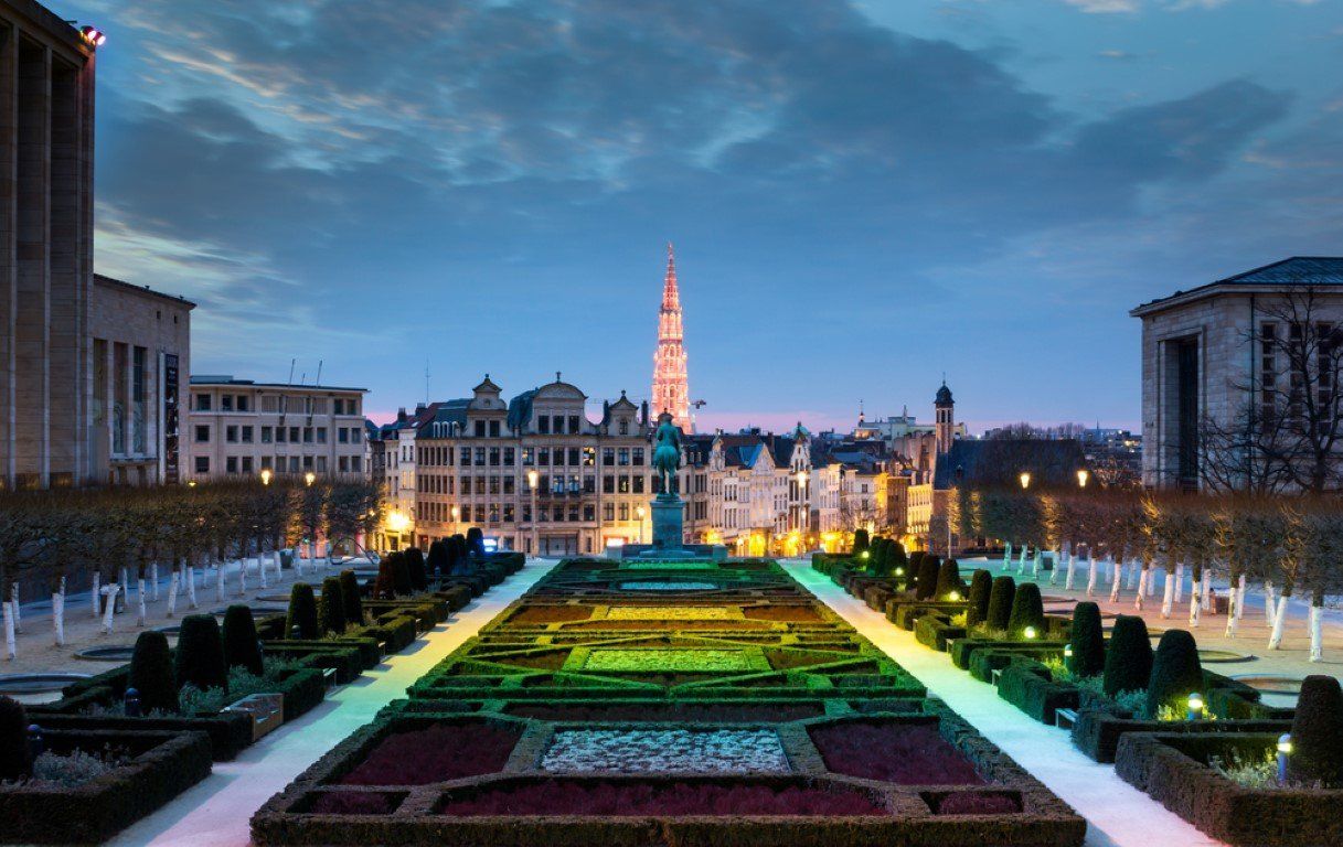 Park with flower beds and statue, buildings in the background, and a tall tower at twilight in Brussels, Belgium.