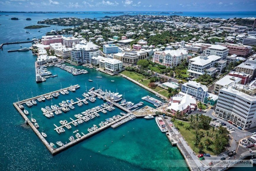 Aerial view of a harbor with boats docked, surrounded by white buildings and a town, all under a blue sky.