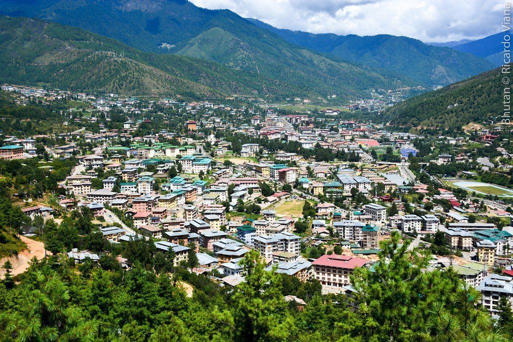Panoramic view of Thimphu, Bhutan, nestled in a valley surrounded by forested mountains, with buildings and greenery.