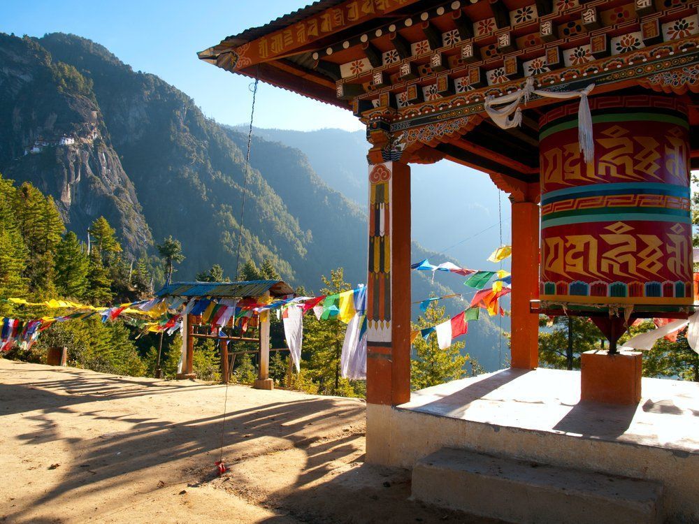 A Buddhist prayer wheel in a wooden structure, colorful flags, and mountains in the background.
