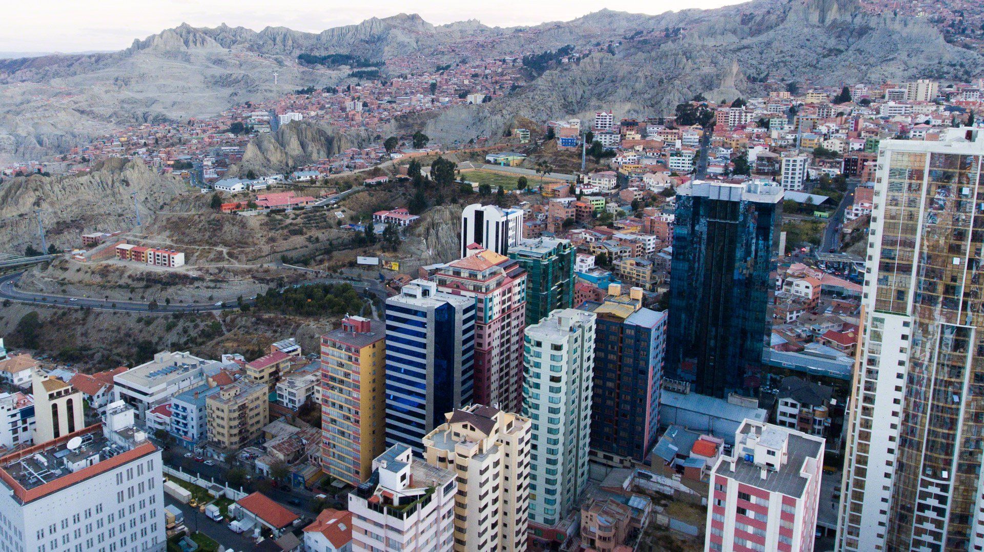 Cityscape with tall buildings in the foreground, mountains and a sprawling city in the background. Overcast sky.