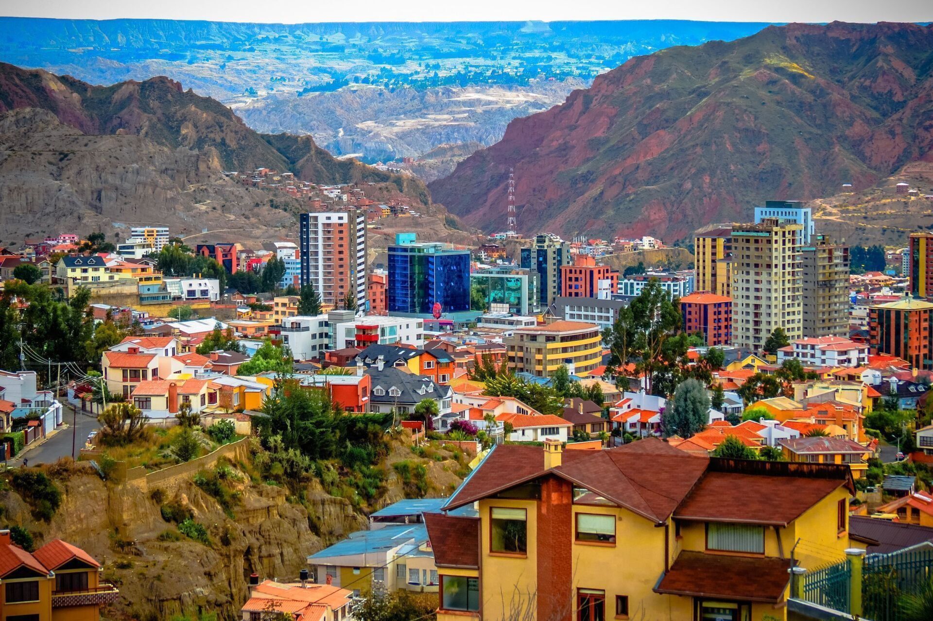 Cityscape of La Paz, Bolivia, nestled in a valley surrounded by reddish-brown mountains. Buildings of various heights and colors dot the landscape.