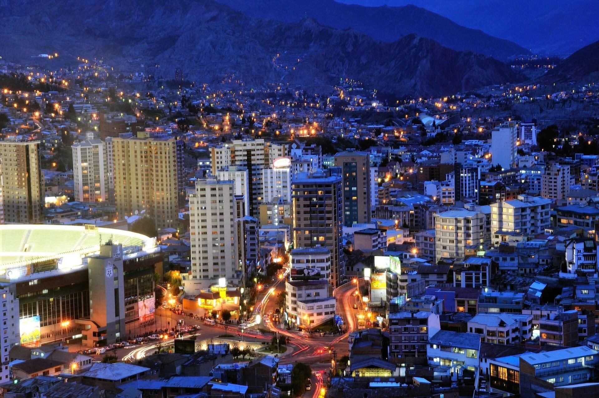 City skyline at dusk, featuring illuminated high-rise buildings against a dark blue mountain backdrop.