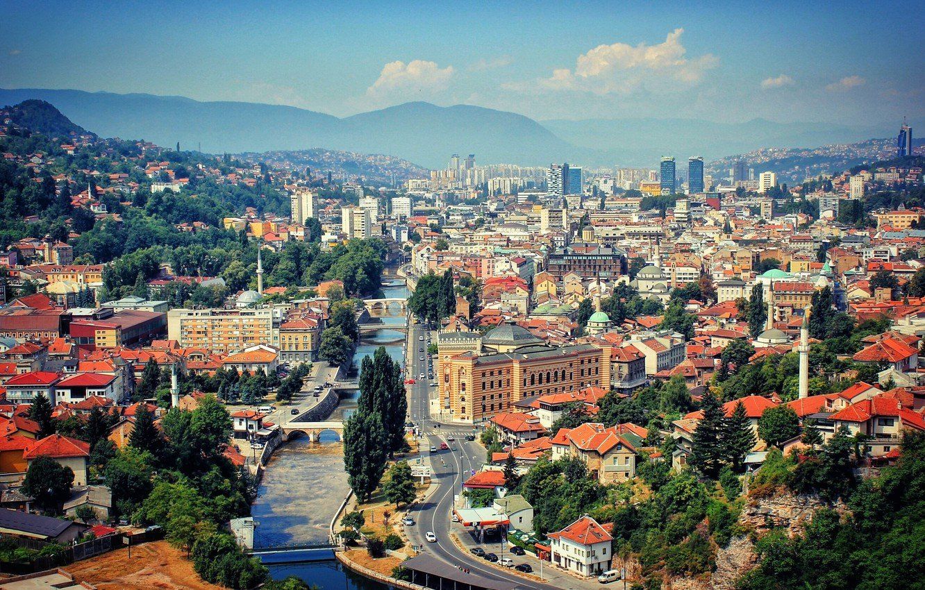 Panoramic view of Sarajevo, Bosnia, with buildings, river, and green hills under a blue sky.