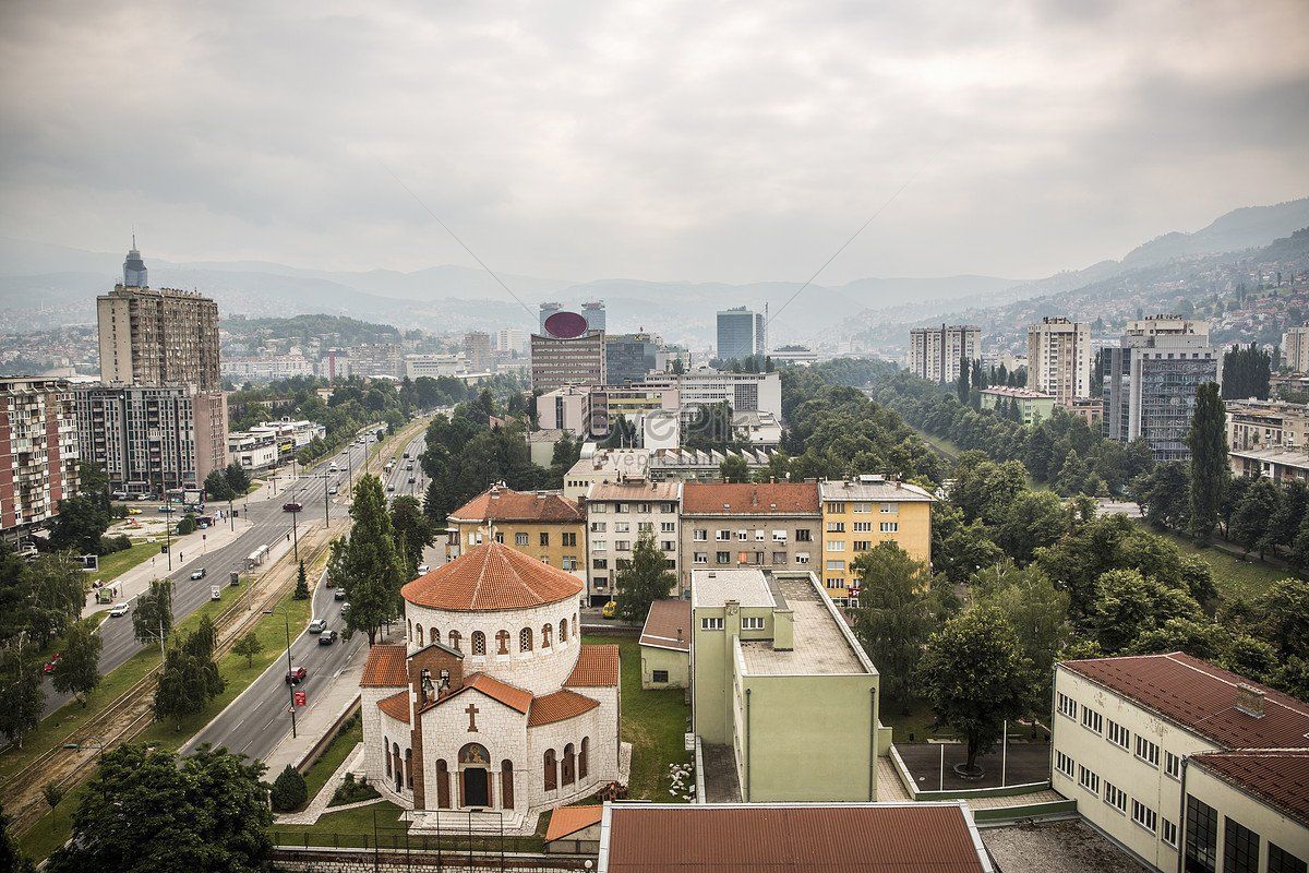 Overhead view of Sarajevo, Bosnia and Herzegovina.  A church with a red-tiled roof sits in the foreground with city buildings and mountains in the background.