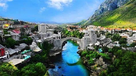 A stone bridge spans a turquoise river in Mostar, Bosnia and Herzegovina, with buildings and mountains in the background.