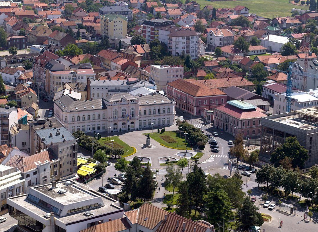 Aerial view of a European city square with ornate buildings, trees, and a monument, under a bright sunny sky.