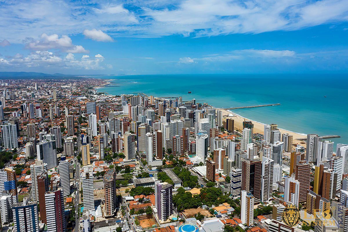 Aerial view of a bustling coastal city, with numerous high-rise buildings and a sandy beach bordering the turquoise ocean under a bright blue sky.