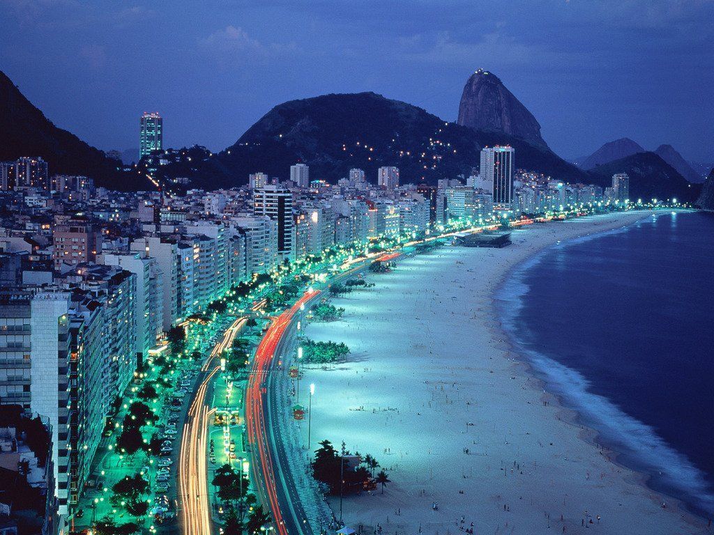 Night view of Copacabana Beach in Rio de Janeiro, Brazil. Illuminated buildings line the beach with Sugarloaf Mountain in the background.