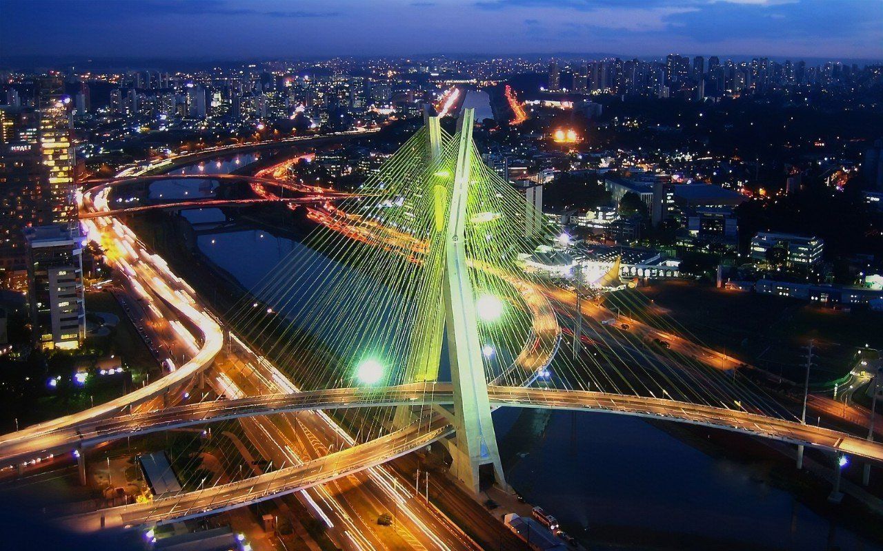 Illuminated cable-stayed bridge at night in São Paulo, Brazil, with city lights and flowing car lights across the water.