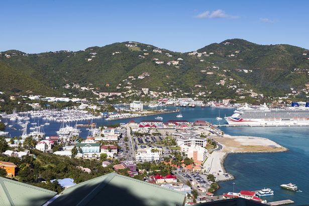 A harbor town nestled at the foot of green hills, with boats, buildings, and a cruise ship docked.