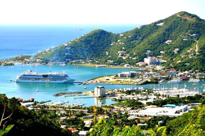A cruise ship docked in a harbor surrounded by green hills and a coastal town on a sunny day.