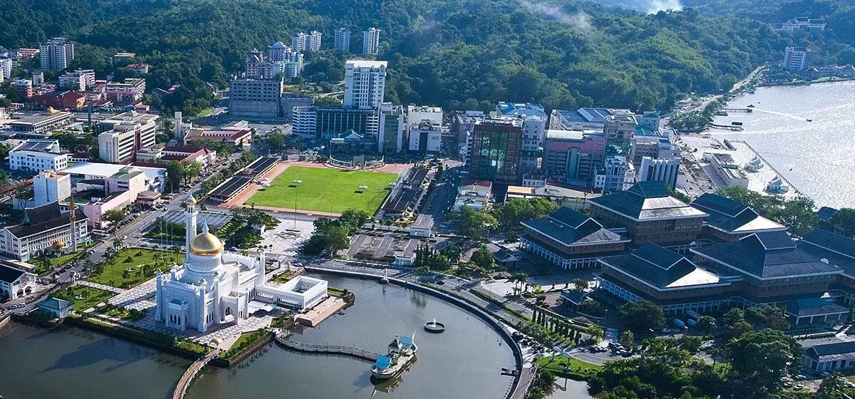 Aerial view of a coastal city with a white mosque, green field, and buildings. Blue water and forested hills are in the background.