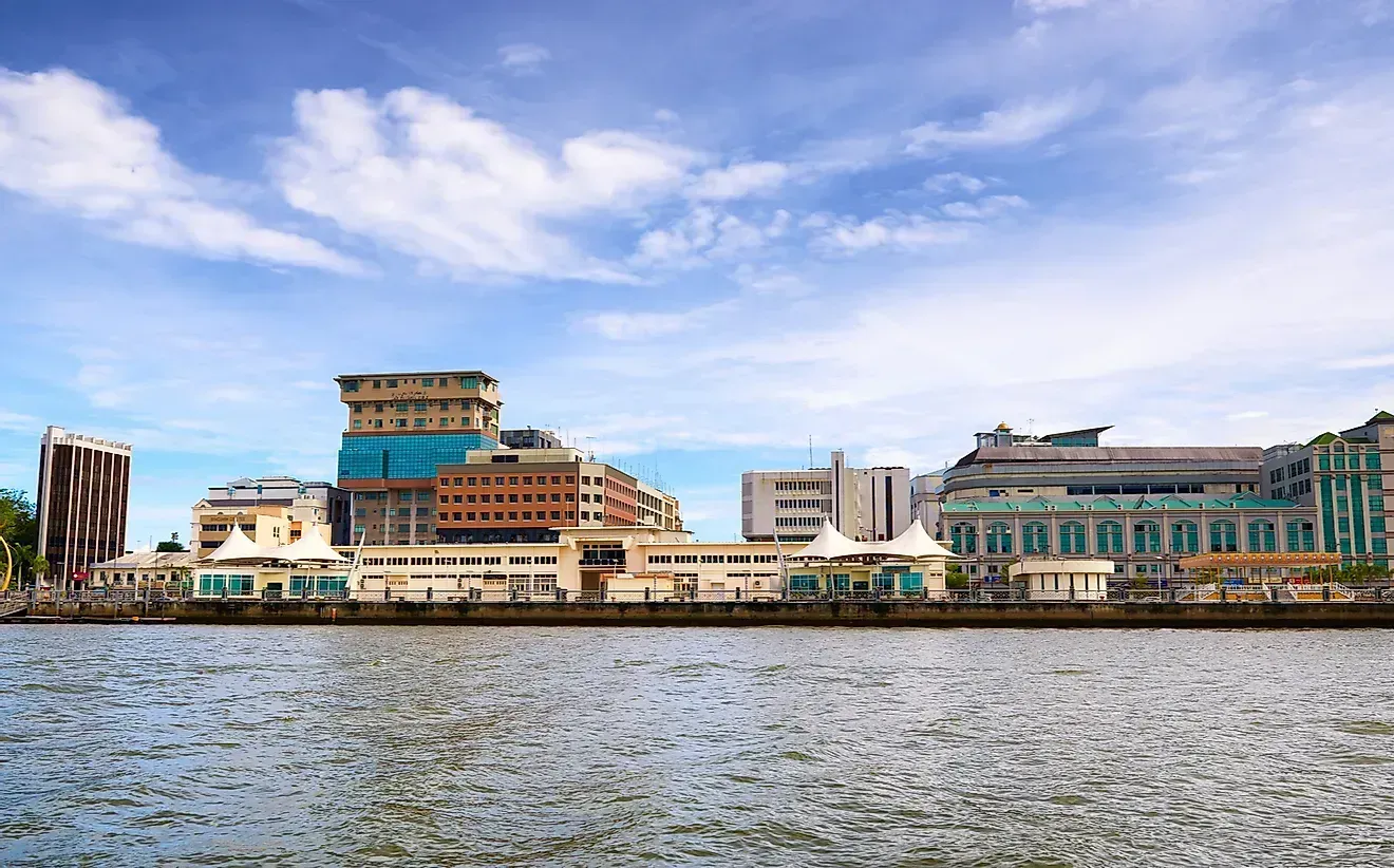 Skyline of buildings along a body of water, under a blue sky with scattered clouds. Buildings vary in height and design, in tones of beige, brown, and white.
