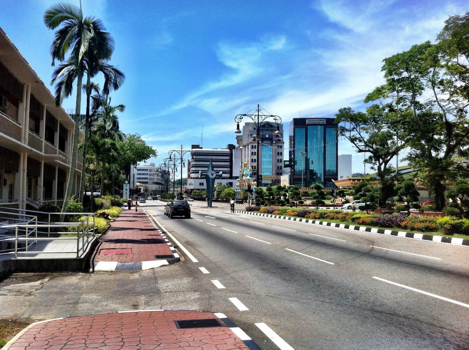 Street view with buildings, palm trees, and blue sky. A car drives on the road lined with a sidewalk.