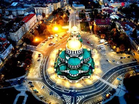 Aerial night view of Alexander Nevsky Cathedral in Sofia, Bulgaria, illuminated with green domes and surrounded by well-lit roads and buildings.