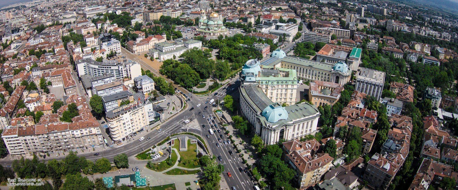Aerial view of Sofia, Bulgaria, showing urban architecture with several buildings with green domes and dense green spaces.