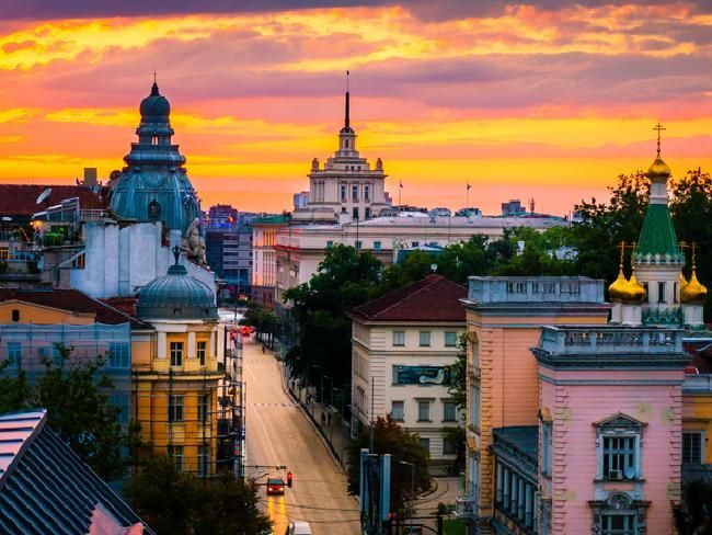 Cityscape of Sofia, Bulgaria at sunset; buildings in various architectural styles under an orange and yellow sky.