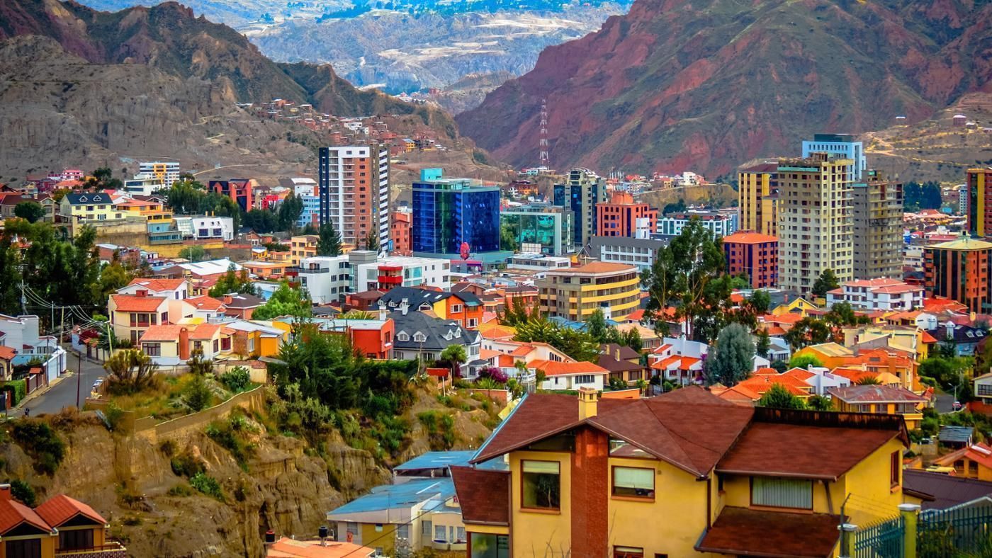 Cityscape of La Paz, Bolivia, with colorful buildings nestled in a valley surrounded by red and gray mountains under a cloudy sky.