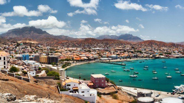 Coastal city with boats on turquoise water, brown hills, and a blue sky dotted with clouds.