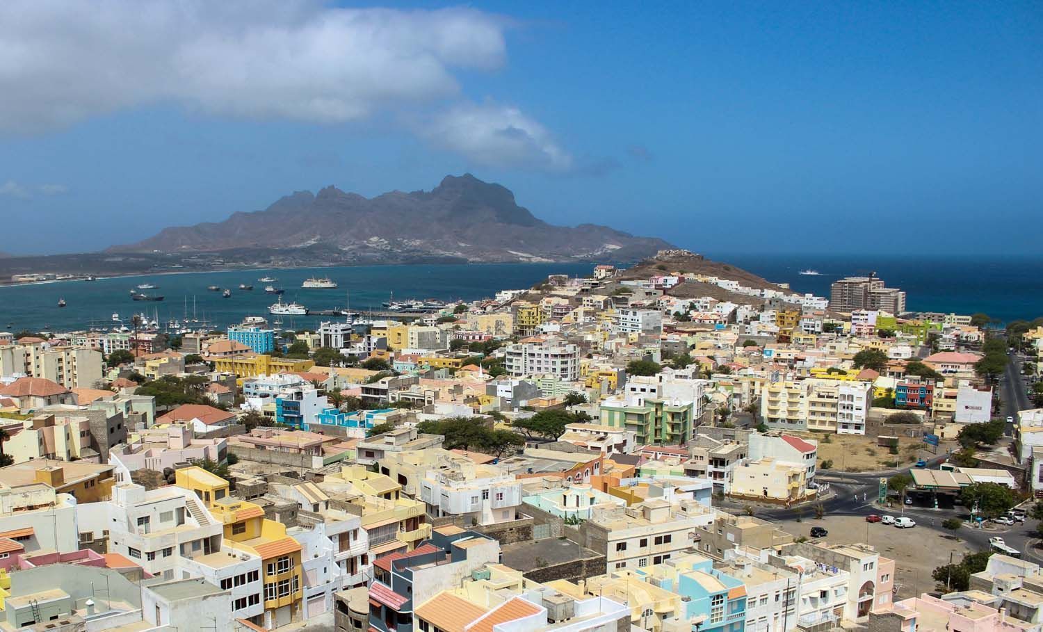 Cityscape of Mindelo, Cape Verde, with colorful buildings, a harbor with boats, and a mountain in the background under a blue sky.