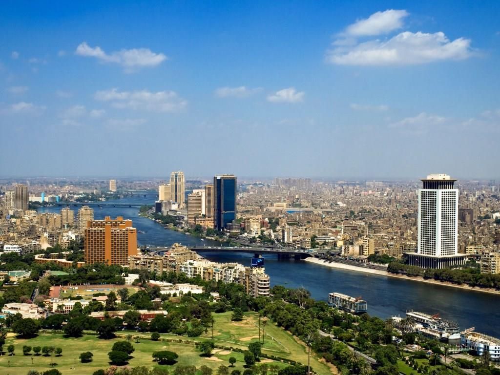 Aerial view of Cairo, Egypt, with the Nile River running through the city. Buildings line the riverbanks under a blue sky with clouds.