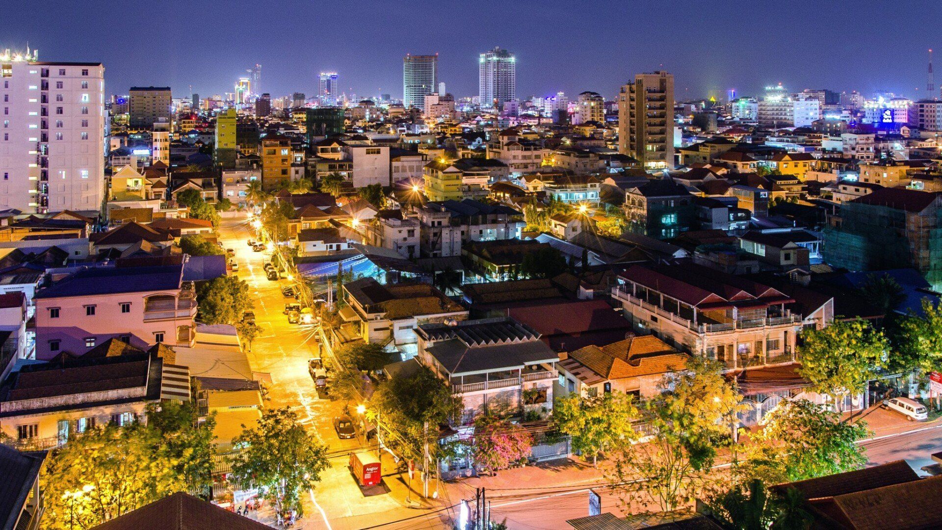 Nighttime cityscape of Phnom Penh, Cambodia, with illuminated buildings, roads, and streetlights. Tall buildings punctuate the skyline.