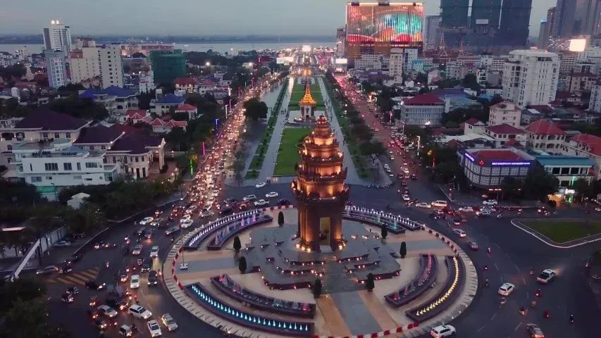Aerial view of Phnom Penh, Cambodia, at dusk. The Independence Monument is at the center of a busy traffic circle, surrounded by city buildings and illuminated roads.