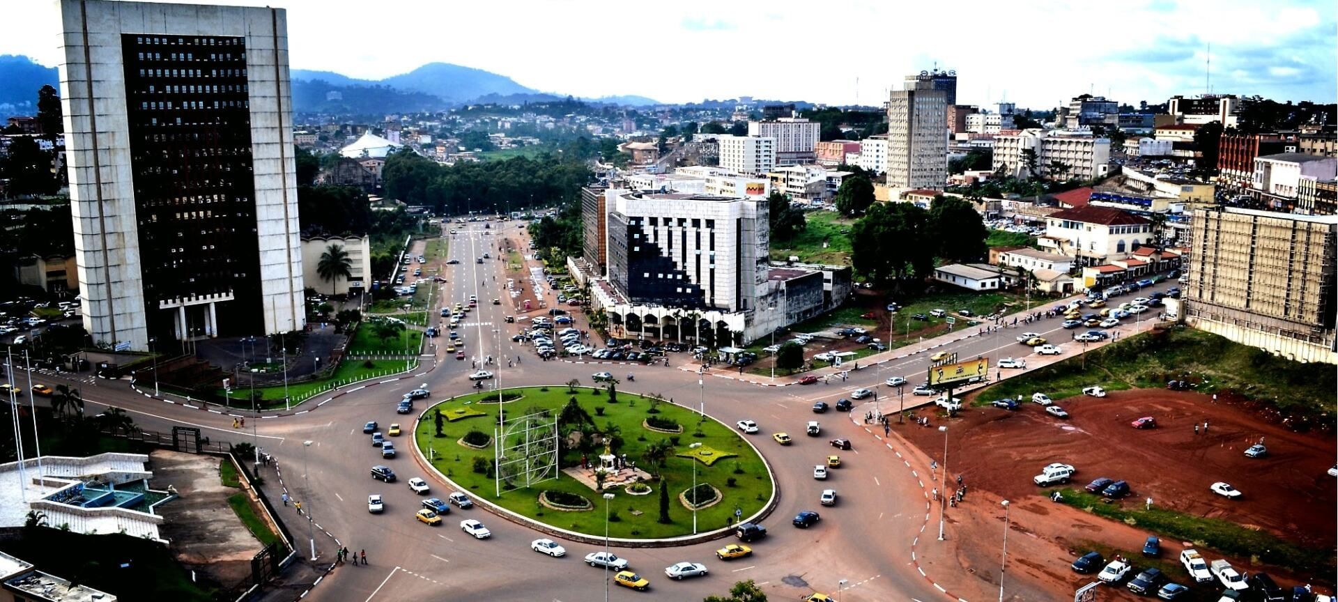 Aerial view of Yaoundé, Cameroon, with skyscrapers, a central roundabout, and busy roads with car traffic.
