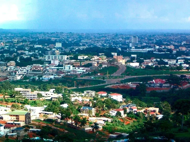 A wide aerial view of Yaoundé, Cameroon. The city sprawls with buildings of varied heights, connected by roads, with green hills in the foreground.