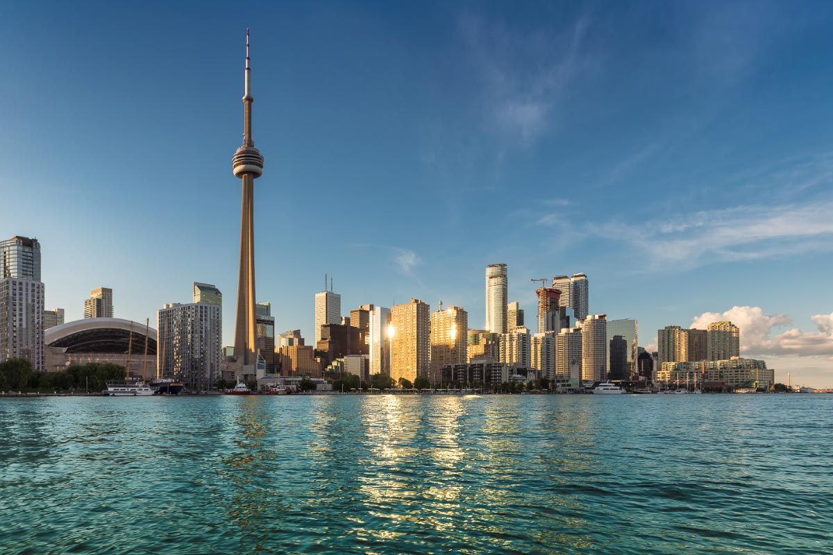 Toronto skyline with the CN Tower, buildings, and reflecting sunlight on the water. Blue sky and water in the foreground.