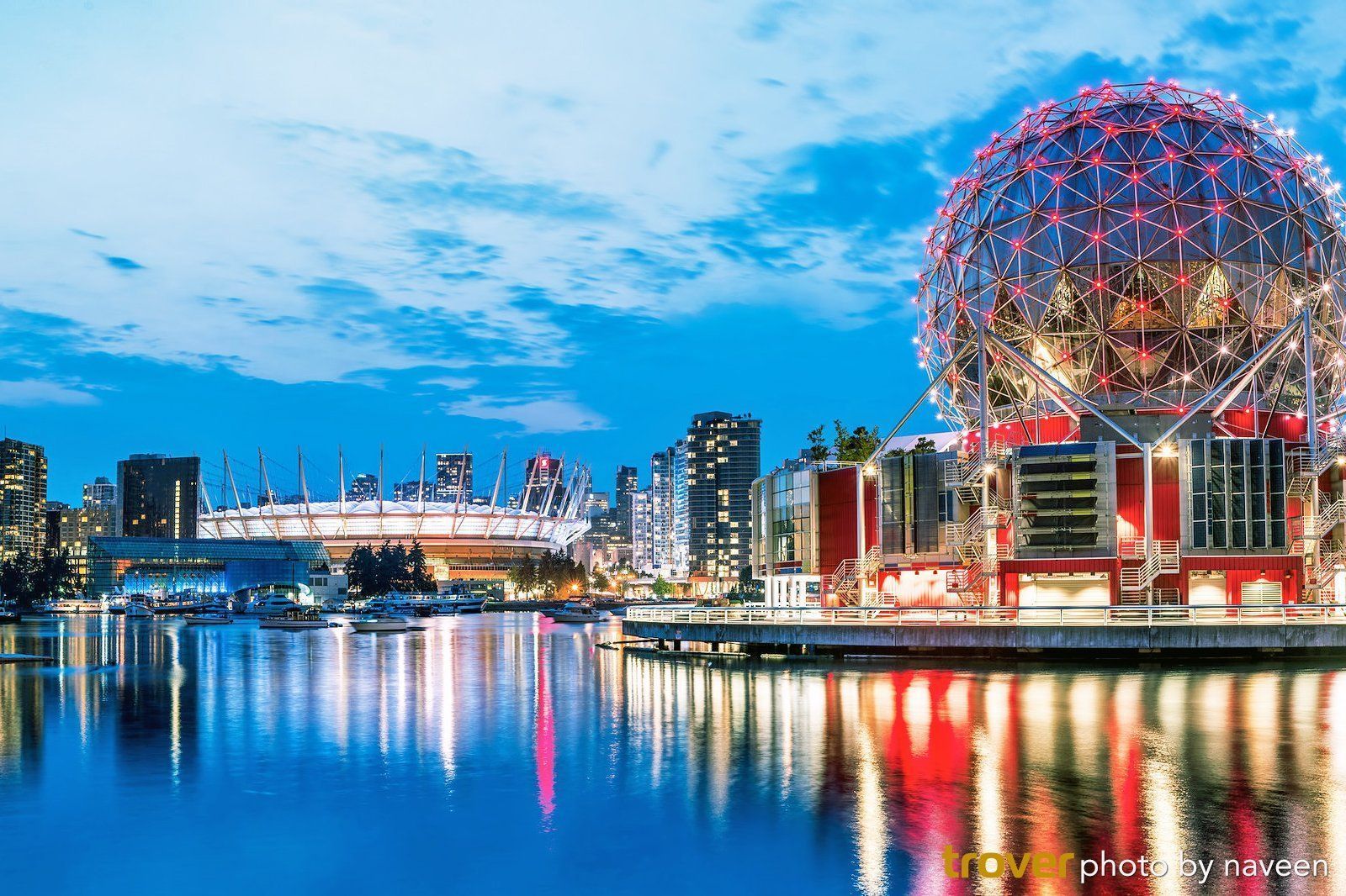 A cityscape at dusk with a glowing geodesic dome and stadium reflecting in the water.