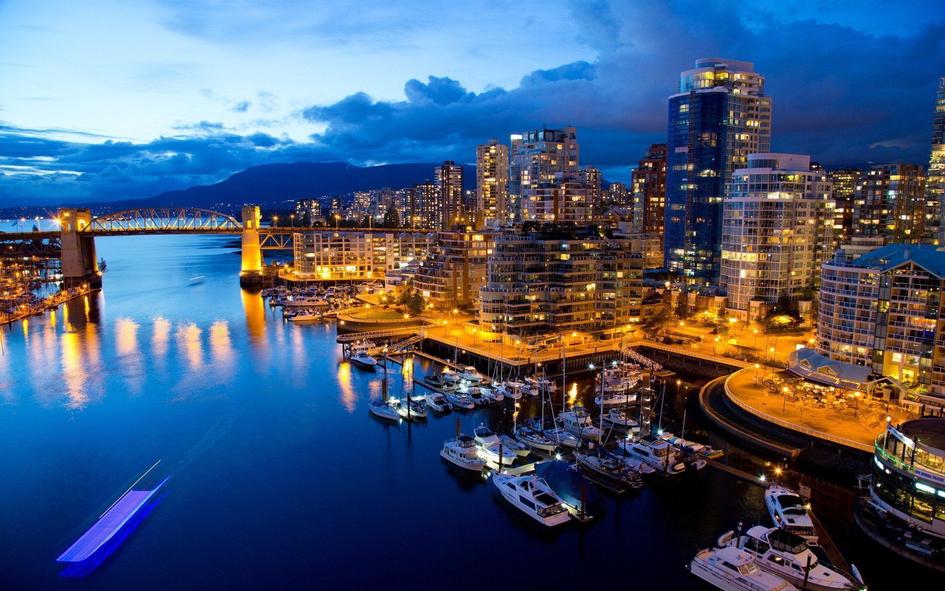 Night view of Vancouver, Canada, with city skyline, harbor, and boats under a blue and gold sky.