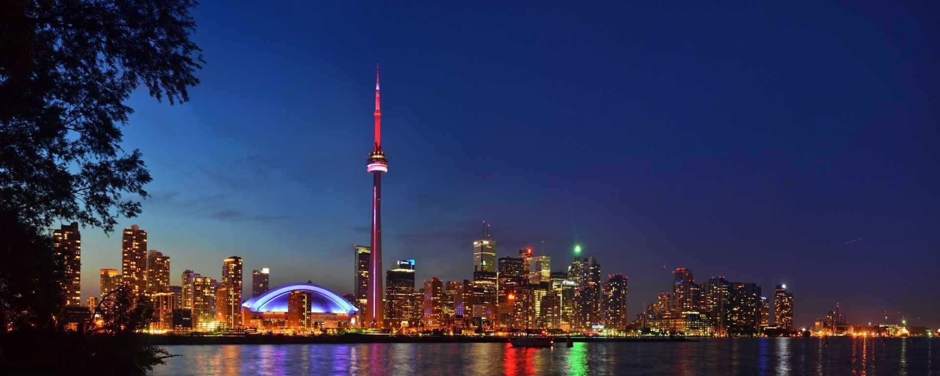Night view of Toronto skyline, featuring the CN Tower, illuminated buildings, and the Rogers Centre, reflecting on the water.