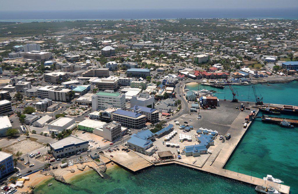 Aerial view of George Town, Grand Cayman, with a harbor, blue water, buildings, and docks under a bright sky.