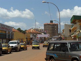 Street scene in Banjul, Gambia, with cars and taxis on a red dirt road, buildings along the sides, and blue sky above.