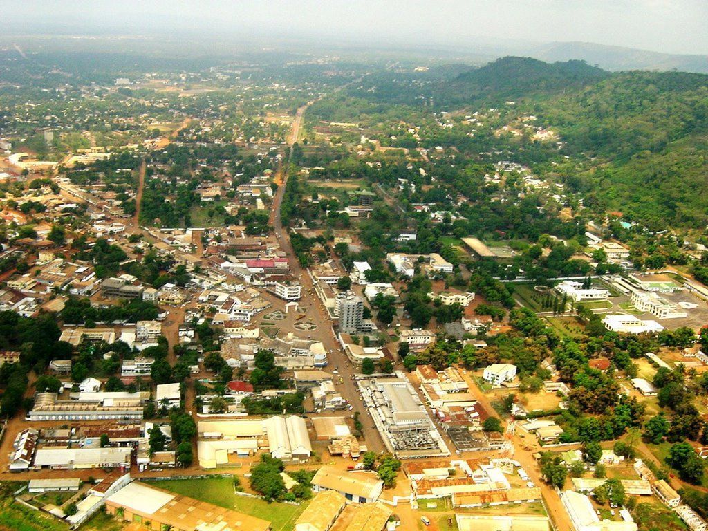 Aerial view of a town with buildings lining a central road, surrounded by green trees and hills.