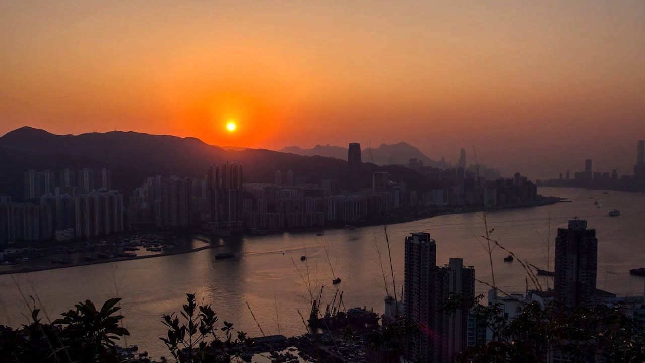 Sunset over a city skyline, with buildings lining a river. The sun is a bright orange orb.