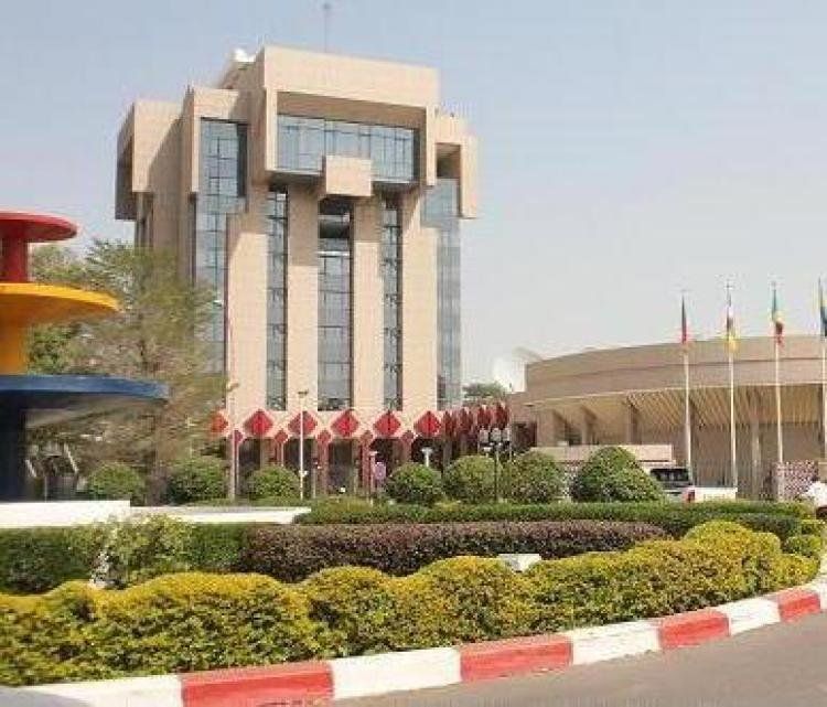 A tall, modern building with many windows, flags, and a landscaped front yard, likely a government or institutional office in an urban setting.