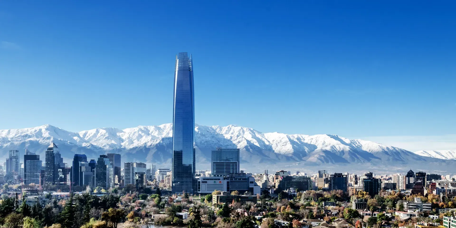 A tall skyscraper dominates the Santiago, Chile skyline, with snow-capped Andes mountains in the background under a clear blue sky.