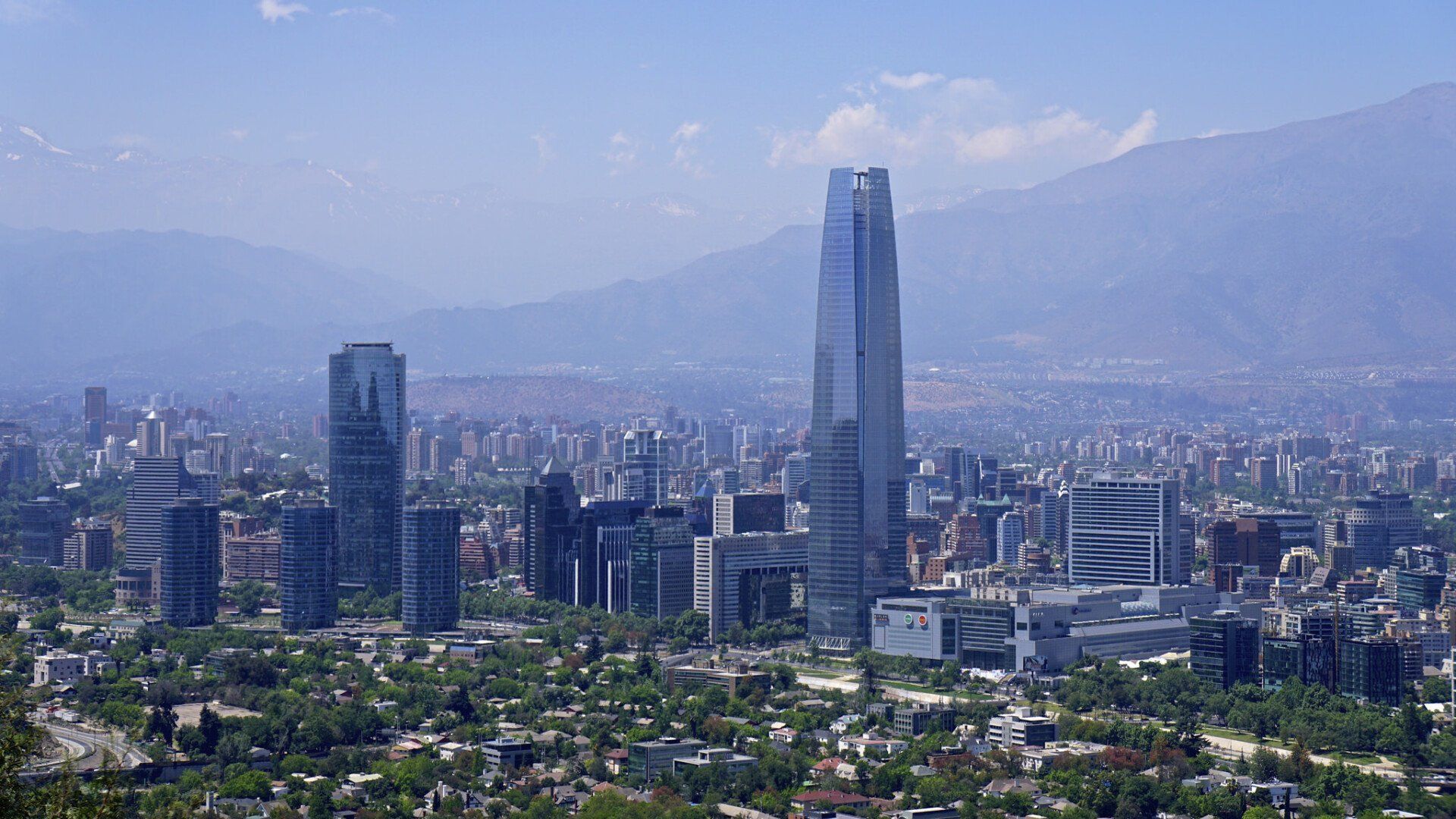 Santiago, Chile cityscape dominated by a tall, modern skyscraper, with the Andes Mountains in the background under a clear sky.