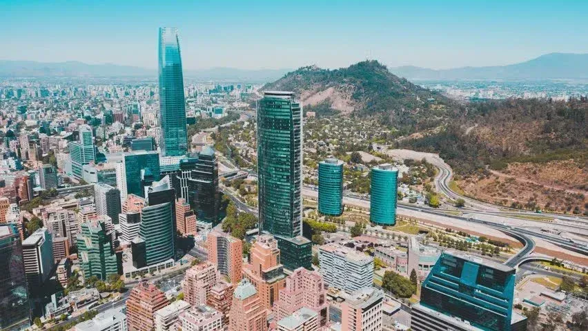 Aerial view of Santiago, Chile, featuring skyscrapers in the city center against a backdrop of hills under a blue sky.