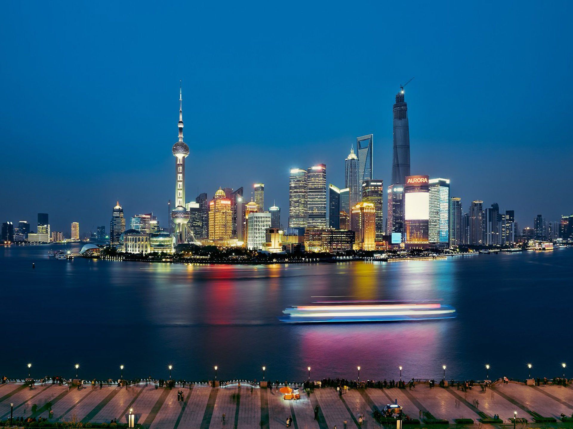 Nighttime skyline of Shanghai, China, with brightly lit skyscrapers and a boat on the water; a dark blue sky provides the backdrop.