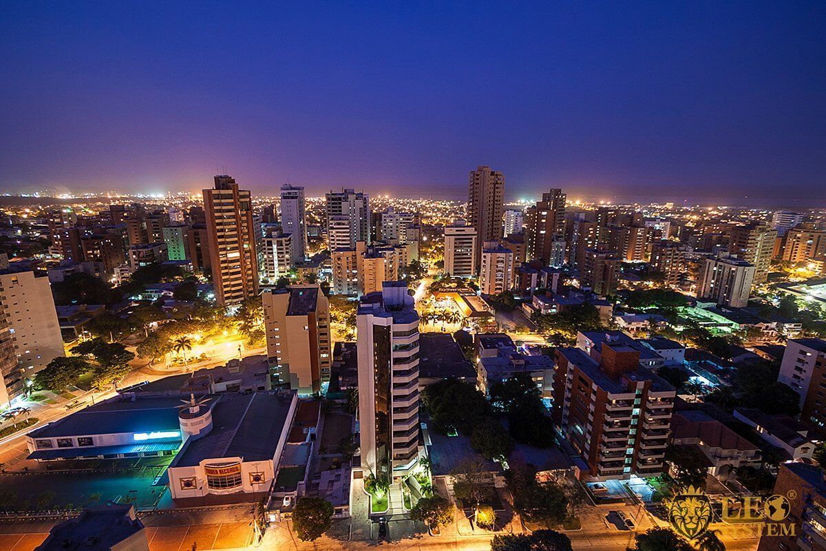 City skyline at night, featuring illuminated buildings under a deep blue sky. Streetlights and building lights create a warm glow.