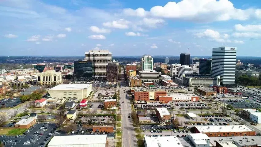 Aerial view of a downtown cityscape with tall buildings, a central road, and a bright blue sky with clouds.