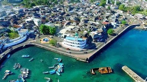 Aerial view of a coastal town with a white building and boats in turquoise water. Buildings crowd the land.